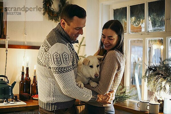 Junges glückliches Paar  Mann und Frau  die einen süßen weißen Hund halten und sich umarmen  während sie in der weihnachtlich geschmückten Küche stehen  schöne Familie mit Welpe  die die Winterferien zusammen in einem gemütlichen warmen Haus genießen