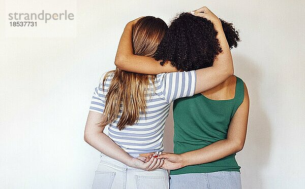 Starke weibliche Freundschaft. Rückansicht zwei Teenagermädchen beste Freunde halten Hände hinter dem Rücken und umarmen  während vor beige Wand stehen