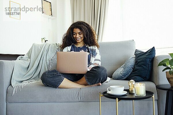 Aufgeregte  fröhliche junge schwarze Frau  die zu Hause auf dem Sofa einen Laptop benutzt  gute Nachrichten erhält  Freude empfindet  Hausaufgaben macht oder online arbeitet  lacht  sich über einen Preis freut