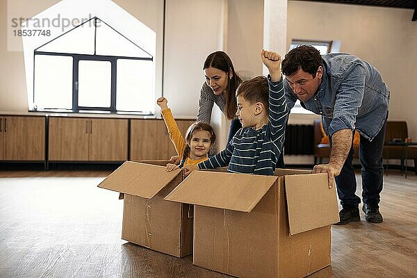 Glückliche Eltern mit Kindern im Wohnzimmer  zwei niedliche Kinder  Junge und Mädchen  sitzen lachend in Kartons und schieben Mutter und Vater. Die Familie feiert den Umzugstag