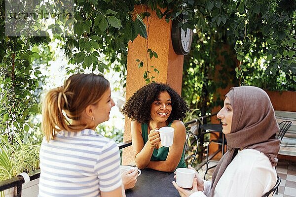 Multiethnische Gruppe von Mädchen in Freizeitkleidung und traditionellem Hijab  die sich im Freien unterhalten und Spaß haben. Drei junge Teenagermädchen in einem Gartencafé trinken Tee oder Kaffee