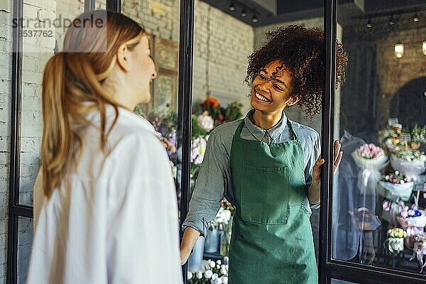 Glückliche schwarze Unternehmerin steht in einem Blumenladen und verkauft frische Blumen an Kunden. Junge blonde Mädchen kaufen einen frischen Blumenstrauß von Florist. Lächelnde afrikanische Frau Botaniker  Verkauf von Blumen