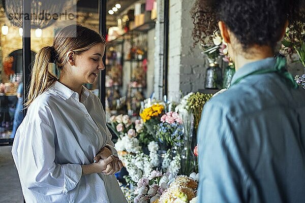 Glückliche schwarze Unternehmerin steht in einem Blumenladen und verkauft frische Blumen an Kunden. Junge blonde Mädchen kaufen einen frischen Blumenstrauß von Florist. Lächelnde afrikanische Frau Botaniker  Verkauf von Blumen