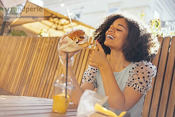 Junges afrikanisches amerikanisches Mädchen ißt klassischen Burger und trinkt Limonade in der Stadt. Outdoor Frau trägt Sommerbluse mit Tupfen eatting leckeren Burger beim Sitzen auf der Terrasse. Frau in blau Verkostung Sandwich mit freudigen Blick bewundern sonnigen w