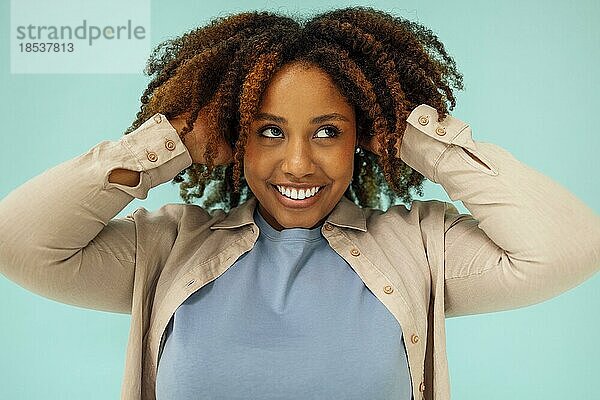 Positives afrikanisches amerikanisches weibliches Modell  das in die Kamera lächelt und sauberes lockiges Haar nach der Hygienebehandlung auf blauem Hintergrund berührt