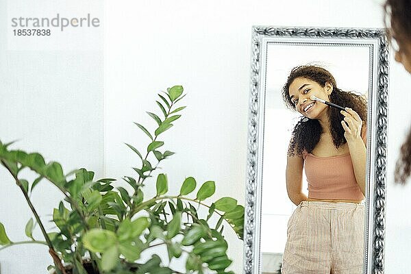 Fröhliche  fröhliche afroamerikanische junge Frau  die Make up mit einem Pinsel aufträgt  vor einem großen Spiegel zu Hause steht  einen Pyjama trägt und ihr eigenes Spiegelbild mit einem breiten Lächeln betrachtet. Schönheit Routine Konzept
