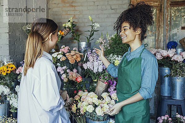 Glückliche schwarze Unternehmerin steht in einem Blumenladen und verkauft frische Blumen an Kunden. Junge blonde Mädchen kaufen einen frischen Blumenstrauß von Florist. Lächelnde afrikanische Frau Botaniker  Verkauf von Blumen