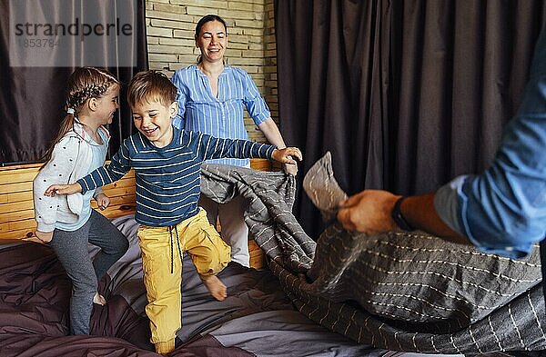 Glückliche Familie multiethnische Mutter  Vater und Kinder  Sohn und Tochter  lachend  spielend  kämpft Kissen und springen im Bett im Schlafzimmer zu Hause am Morgen