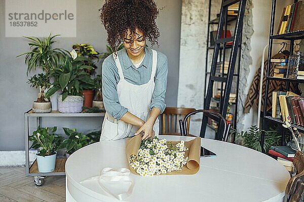 Zarte Verkäuferin  die ihre Arbeit genießt  während sie einen Blumenstrauß zum Verkauf anbietet. Junge Floristin in Schürze  die einen schönen einfachen Gänseblümchenstrauß in Bastelpapier bindet. Porträt eines lächelnden Mädchens mit dunklen lockigen Haaren  das mit Freude auf einen Kamillenstrauß blickt