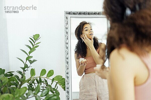 Hautpflege-Routine. Afro Girl Applying Eye Cream Looking In Mirror Standing In living room. Selektiver Fokus. Gesichtspflege