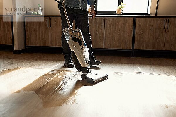 Brünetter Mann saugt mit Staubsauger den Holzboden im Loft Wohnzimmer. Nahaufnahme