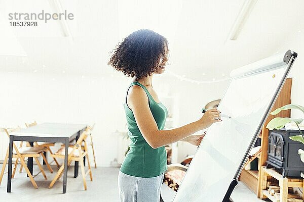 Schönes afroamerikanisches Mädchen mit dunklen lockigen Haaren steht in der Nähe von Brett und gibt Präsentation an Kollegen im Büro. Junge Geschäftsfrau schaut auf ihre Mitarbeiter  während sie ein neues Projekt bespricht