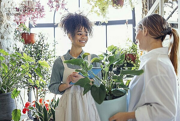 Glückliche schwarze Unternehmerin steht in einem Blumenladen und verkauft frische Blumen an Kunden. Junge blonde Mädchen kaufen einen frischen Blumenstrauß von Florist. Lächelnde afrikanische Frau Botaniker  Verkauf von Blumen