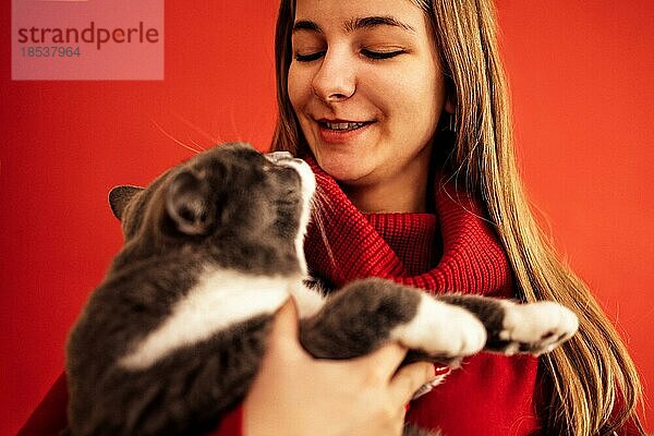 Porträt eines blonden Teenagers in einem roten Pullover  der eine Katze auf der Hand hält. Schönes lächelndes Mädchen mit ihrem Haustier. Nahaufnahme. Raum kopieren. vor einem roten Hintergrund