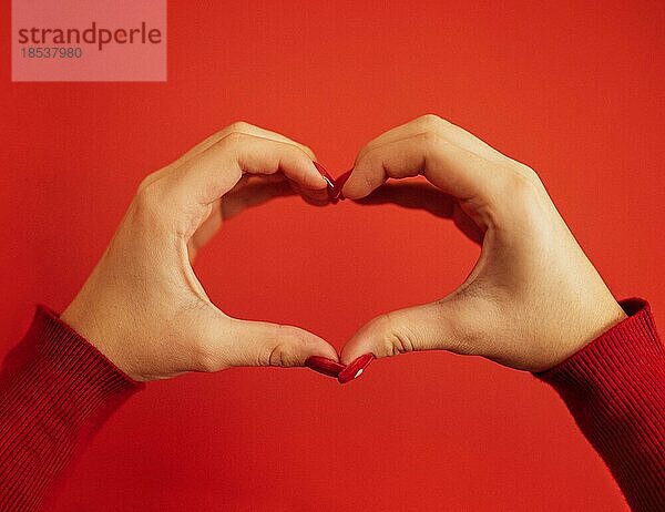 Weibliche Hände bilden eine Herzform. Hände eines jungen weißen Mädchens mit einer schönen leuchtend roten Maniküre. Nahaufnahme. Valentinstag Konzept. Roter Hintergrund