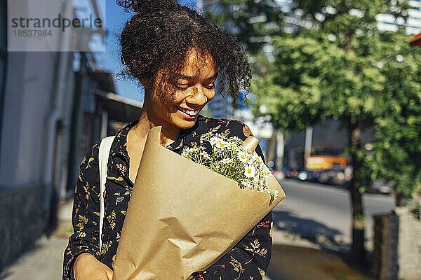 Junge afroamerikanische Frau hält Blumen und lächelt. Glückliche schöne dunkelhäutige Mädchen geht entlang der Straße mit einem Blumenstrauß von Gänseblümchen. Positive Modell im Freien auf der Stadt Sommer Hintergrund. Kopieren Raum