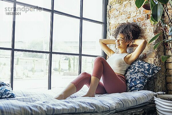 Zufriedenes entspanntes afroamerikanisches Mädchen in Hauskleidung auf der Fensterbank sitzend  glückliche afroamerikanische Frau zu Hause  die sanft lächelnd zum Fenster schaut