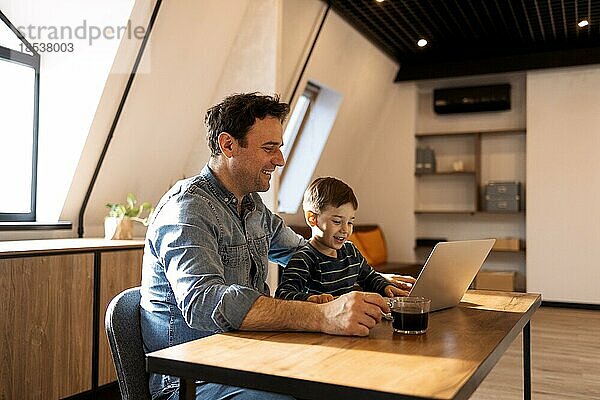 Brunette Vater und Sohn mit einem Laptop  während Vater trinken Frühstück Kaffee  sie tragen legere Kleidung in gemütlichen Loft Hintergrund