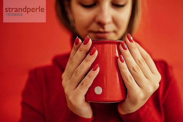 Junges blondes Mädchen in einem roten Pullover hält eine rote Tasse in den Händen. Nahaufnahme einer schönen leuchtend roten Maniküre. Valentinstag Konzept. Kaffeepause. Zeit zum Entspannen