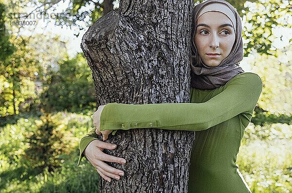Muslimisches Teenagermädchen umarmt einen Baum. Close up Porträt einer jungen Frau in einem Hijab und Freizeitkleidung im Park. Schutz der Umwelt und Pflege von Pflanzen Konzept