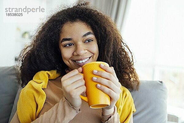 Fröhlich lächelndes afrikanisches Mädchen  das sich auf der Couch mit einer Tasse Kaffee ausruht  zu Hause ausruht  den freien Tag genießt  warme Kleidung trägt