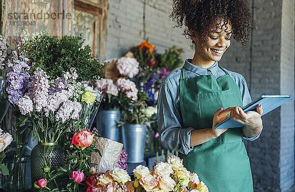 Junge schwarze Frau mit beiger Schürze auf Blumenladen Hintergrund mit Kopierraum. Porträt einer erfolgreichen afroamerikanischen Frau  die auf einen Tabletcomputer schaut