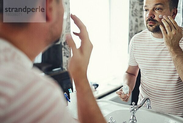 Millennial gut aussehend Mann in den Spiegel schauen  Anwendung feuchtigkeitsspendende nach Rasierschaum auf Wangen im Bad  Kopfschuss Nahaufnahme. Gepflegter junger Mann bei der morgendlichen Hautpflege nach dem Duschen