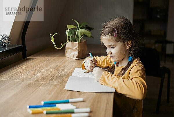 Nettes brünettes kleines Mädchen  das am Küchentisch sitzend etwas in ihr Notizbuch schreibt