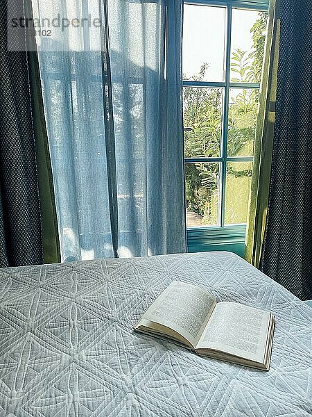 Ein aufgeschlagenes Buch liegt auf einer weißen Steppdecke auf dem Bett. Das Sonnenlicht scheint und der grüne Garten ist durch die Schlafzimmerfenster sichtbar. Gemütliches Interieur. Zeit für sommerliche Ruhe und Entspannung