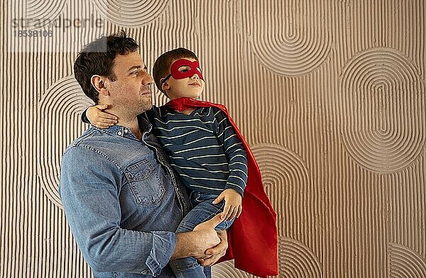 Joyful Papa trägt kleines Kind  das ihn umarmt und lächelt glücklich  in Superhelden Kostüme gekleidet  vorbeige Hintergrund  haben Spaß zusammen. Väter Tag Konzept