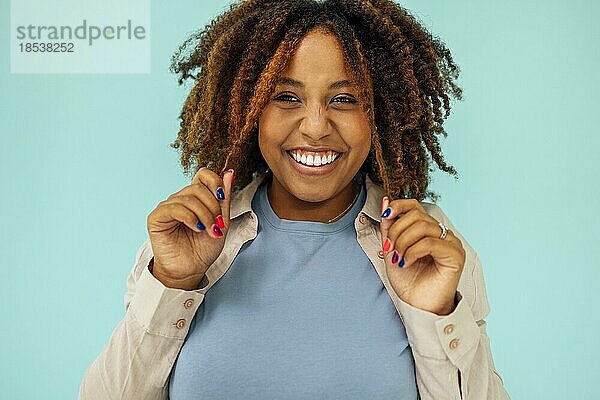 Positives afrikanisches amerikanisches weibliches Modell  das in die Kamera lächelt und sauberes lockiges Haar nach der Hygienebehandlung auf blauem Hintergrund berührt