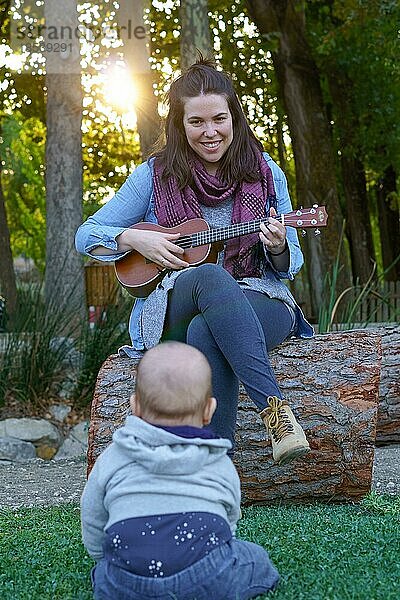 Eine Frau spielt lächelnd Ukulele und ein Baby sitzt im Gras und beobachtet sie als Zuschauer