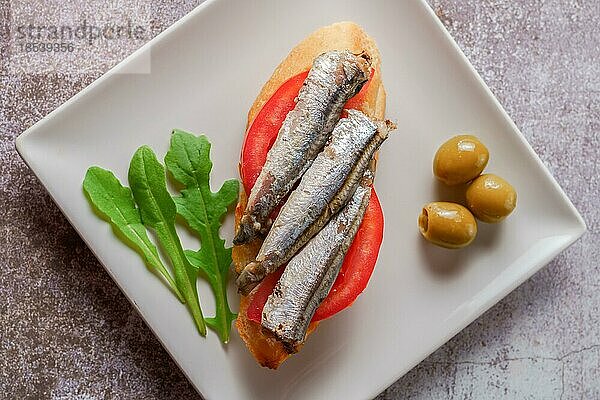 Tapa aus Sardinen auf einer Scheibe Brot mit Tomate und Oliven auf einem weißen Teller mit typisch spanischem weißem Hintergrund