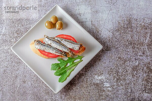 Tapa aus Sardinen auf einer Scheibe Brot mit Tomate und Oliven auf einem weißen Teller mit typisch spanischem weißem Hintergrund