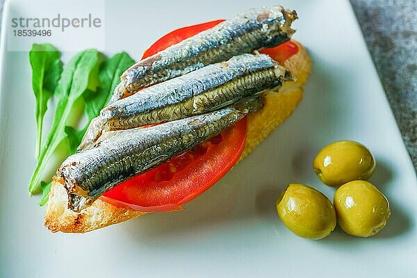 Tapa aus Sardinen auf einer Scheibe Brot mit Tomate und Oliven auf einem weißen Teller mit typisch spanischem weißem Hintergrund