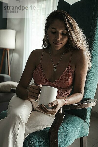 Innenaufnahme einer schönen Latina Frau  die eine Tasse mit heißem Tee oder Kaffee am Morgen hält