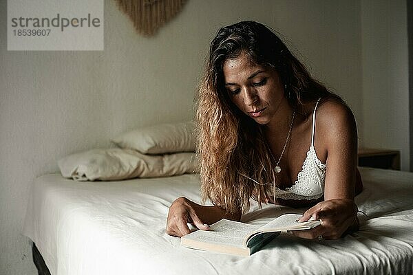 Junges Latina Mädchen mit langen Haaren liest ein Buch auf ihrem Bett in ihrem Zimmer