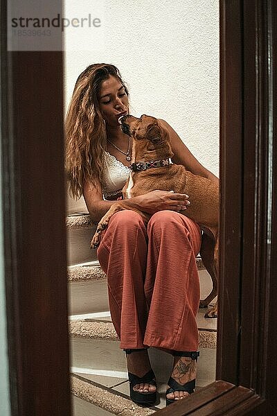 Schöne junge Latina  die ihren Hund umarmt und ihre Zuneigung auf der Treppe ihres Hauses zeigt. Liebe für Tiere