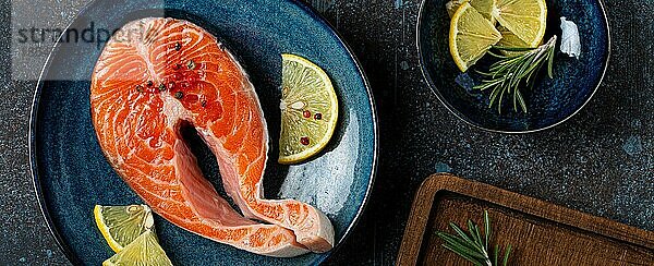 Ungekochte rohen frischen Fisch Lachssteak Draufsicht auf Platte rustikalen dunklen Betonstein Hintergrund mit Rosmarin  Zitrone Keile und Gewürze  Delikatesse gesunden Fisch Kochen und Ernährung Konzept flach legen