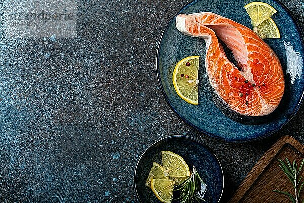 Ungekochte rohen frischen Fisch Lachssteak Draufsicht auf Platte rustikalen dunklen Betonstein Hintergrund mit Rosmarin  Zitronenscheiben und Gewürze  Delikatesse gesunden Fisch Kochen und Ernährung Konzept Raum für Text