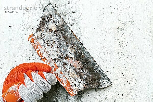 Unkenntlich gemachte Hand eines Stuckateurs hält ein altes Spezialspachtel und kratzt eine Betoninnenwand ab  entfernt alten Putz bei der Renovierung eines Gebäudes. Nahaufnahme  selektiver Fokus auf Arbeitswerkzeug