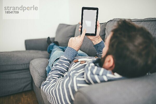 Mann  der ein kleines digitales Tablet oder einen E-Book-Reader benutzt  während er sich auf der Couch entspannt