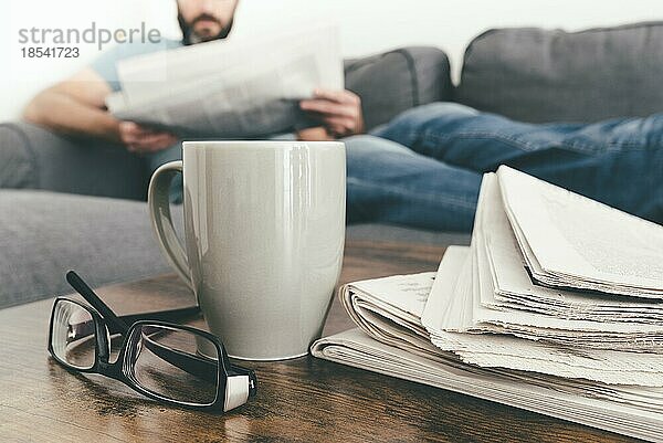 Mann entspannt sich auf dem Sofa und liest eine Zeitung mit einem Stapel Zeitungen und einem Kaffeebecher im Vordergrund