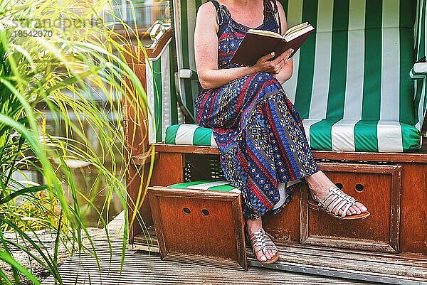 Frau im Sommerkleid entspannt im Strandkorb auf der Terrasse und liest ein Buch