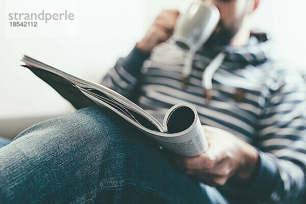 Mann liest Zeitschrift oder Zeitung und trinkt Kaffee  während er sich auf der Couch entspannt