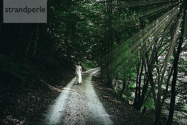 Blonde kaukasische Frau im Sommerkleid geht auf schmalem Schotterweg durch dunklen Wald