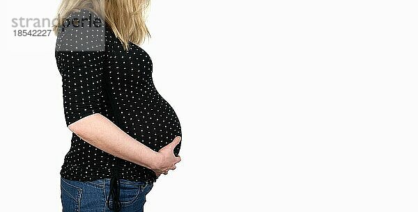Seitenansicht des Mittelteils einer im 9. Monat schwangeren Frau in gepunktetem Hemd und Jeans mit Händen auf dem Bauch vor weißem Hintergrund