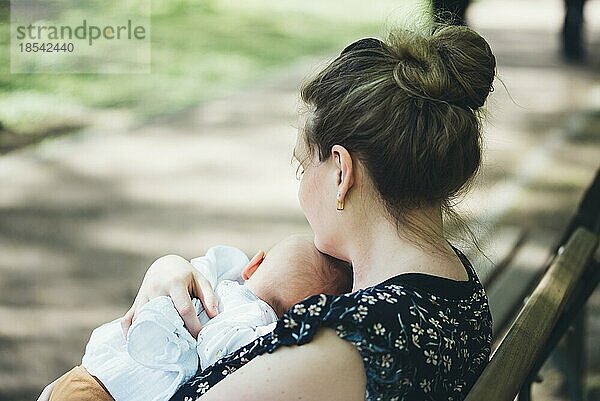 Mutter hält ihr Baby in den Armen auf einer Bank im Park an einem sonnigen Sommertag