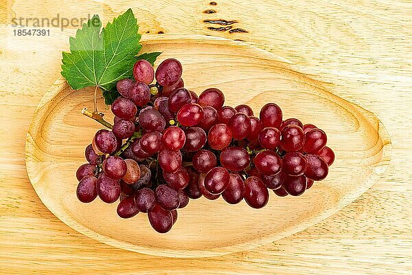 Draufsicht (Flat Lay) auf eine Traube reifer roter Weintrauben mit grünem Blatt in einem Holztablett auf einem Holztisch. Abunch of Crimson Seedless Trauben in einem Holztablett