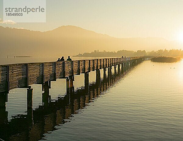 Langer Holzsteg und Promenade über dem Zürichsee bei Rapperswil im goldenen Abendlicht mit Silhouette von Fußgängern und Spaziergängern und Bergen im Hintergrund